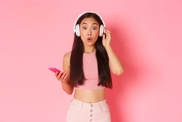 Zaskoczony i podekscytowany azjatycki słodkie dziewczyna gapić się na aparat zastanawiał się podczas słuchania ciekawych podcast lub znajomych playlist, sprawdzanie nowych piosenek w słuchawkach, trzymając telefon komórkowy, różowa ściana - Zdjęcie, obraz