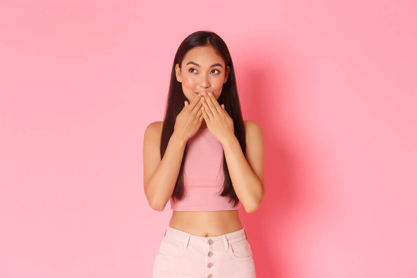 Lifestyle, beauty and women concept. Close-up of dreamy and silly cute asian girl remember romantic moment or kiss, touching lips with hands and looking upper left corner thoughtful, pink background - Photo, Image