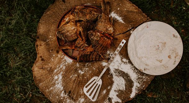 gebakken vis crucian karper ligt op een bord op een oude gebarsten houten stomp waarop witte bloem wordt besprenkeld en een keukenspatel ligt. groen gras. Zomer. picknick. - Foto, afbeelding
