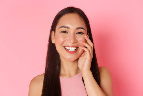Beauty, fashion and lifestyle concept. Close-up of beautiful asian young girl without acne or blemishes, white smile, touching face and looking happy, standing over pink background - Photo, Image