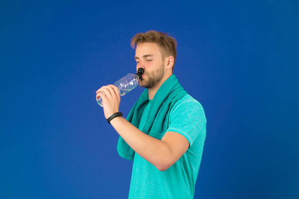 Man on calm face drinking water after training in gym, violet background. Guy with bristle wears fitness tracker. Sporty lifestyle concept. Man with towel on neck renews water balance after training - Photo, image