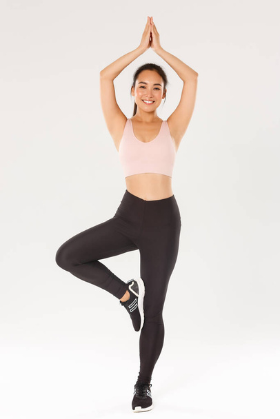 Sport, gym and healthy body concept. Full length of smiling cute sports girl, female athlete in active wear clasp hands above head, meditating, practice yoga, standing in asana pose, white background - Foto, imagen