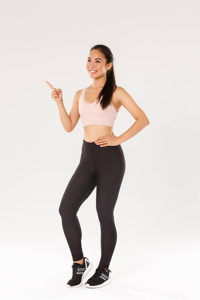 Sport, gym and healthy body concept. Full length smiling satisfied female athlete, asian fitness girl in active wear making her choice, pointing and looking upper left corner, pick gym for training - Fotoğraf, Görsel