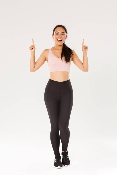 Sport, gym and healthy body concept. Full length of cheerful smiling asian fitness girl, female athelte in sportswear showing advertisement, pointing fingers up and inviting for training, workout - Φωτογραφία, εικόνα