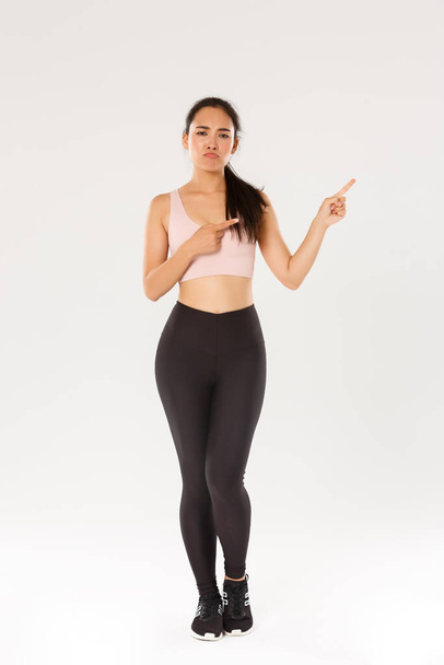Full length of skeptical, disappointed asian fitness girl, sportswoman smirk and sulking while complaining on bad gym or awful workout gear, pointing upper right corner displeased, white background - Foto, afbeelding