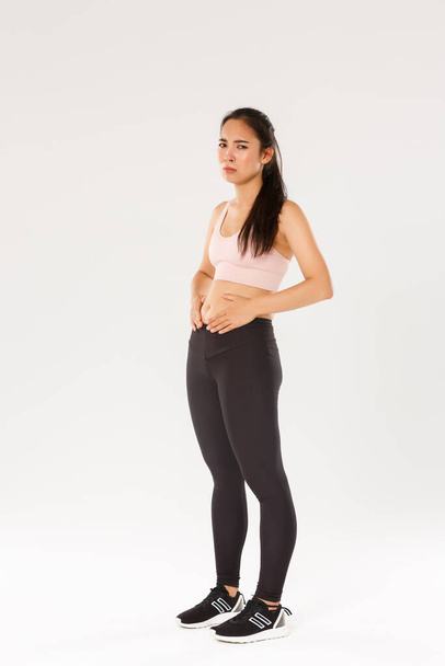 Full length of upset and gloomy asian fitness girl trying to lose weight, touching belly, cant see results from workout or diet, feeling uneasy while standing in sportswear over white background - Photo, Image