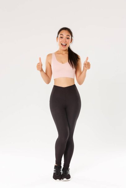 Full length of amazed and satisfied smiling fitness girl, female athelte in active wear liking new gym or workout program, showing thumbs-up pleased, encourage start training or doing exercises - 写真・画像