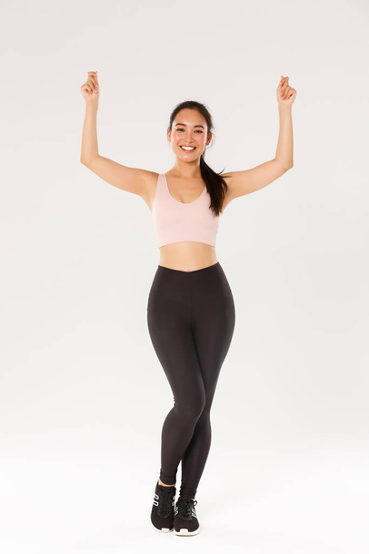 Full length of slim and healthy asian smiling girl workout, standing in sportswear and raising hands up as if holding sign or banner, advertise sports equipment or gym membership discounts - Φωτογραφία, εικόνα