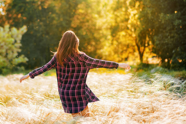 A beautiful young brunette walks in the park, a girl dances and enjoys a beautiful sunset, a woman is dressed in a fashionable plaid dress, a beautiful photo session, free space for text - Photo, Image