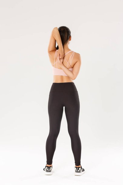 Full length rear view of active and slim brunette asian fitness girl, female athlete warm-up before yoga classes, lock hands behind back, sportswoman doing stretching exercises, white background - Foto, afbeelding