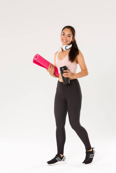 Full length of smiling slim and beautiful asian female athlete, girl carry fitness exercise rubber mat and water bottle, going for yoga classes or workout in gym with own equipment, white background - Photo, Image