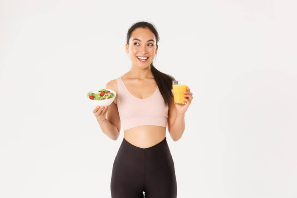 Sport, wellbeing and active lifestyle concept. Smiling fitness girl, asian female athlete looking upper left corner happy, eating healthy salad and orange juice before workout, lose weight with diet - Photo, Image