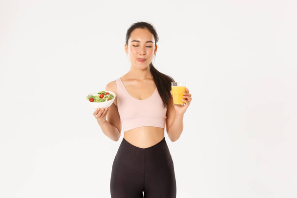Sport, wellbeing and active lifestyle concept. Smiling cute fitness asian girl in activewear, licking lips satisfied with delicious healthy salad and orange juice before workout in gym - Photo, Image
