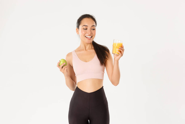 Αθλητισμός, ευεξία και ενεργός τρόπος ζωής. Χαμογελώντας ευτυχισμένη ασιατική fitness girl στο αθλητικό ντύσιμο κοιτάζοντας χυμό πορτοκάλι ευχαριστημένος, τρώγοντας μήλο μετά την παραγωγική προπόνηση στο γυμναστήριο, λευκό φόντο - Φωτογραφία, εικόνα