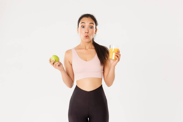 Sport, welzijn en actief lifestyle concept. Amusant en verwonderd schattig aziatisch meisje houdt van fitness en het eten van gezond voedsel, met appel en sinaasappelsap, op zoek verbaasd gezegde wow, witte achtergrond - Foto, afbeelding