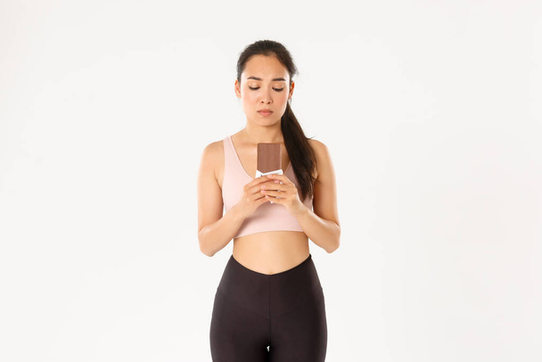 Sport, wellbeing and active lifestyle concept. Indecisive cute asian girl in workout clothing, looking hesitant at chocolate bar, trying resist temptation as losing weight on diet - Foto, imagen