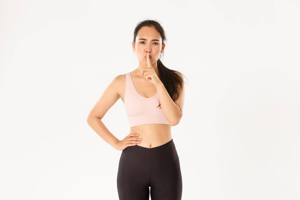 Sport, wellbeing and active lifestyle concept. Serious asian female gym instructor, coach demand discipline in fitness class, shushing with finger pressed to lips, stay quiet, white background - Photo, Image