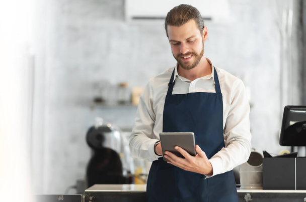Portrait of a Hipster barista in blue apron using digital tablet standing at the bar of the modern cafes. Smiling attractive Bearded man receiving hot drink online orders with service mind - Photo, image