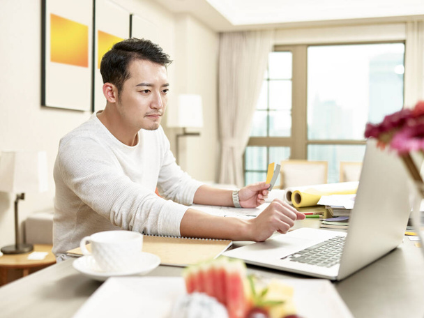 νεαρός Ασιάτης σχεδιαστής επαγγελματίας που εργάζεται από το σπίτι κάθεται στην κουζίνα χρησιμοποιώντας φορητό υπολογιστή (artwork in background digitally modified) - Φωτογραφία, εικόνα