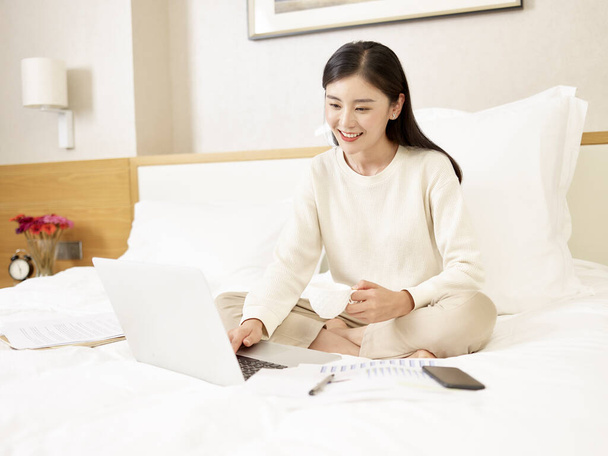 giovane donna d'affari asiatica che lavora da casa seduta sul letto con una tazza di caffè usando il computer portatile - Foto, immagini