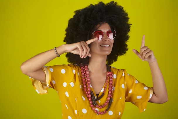 Молодая красивая и улыбающаяся женщина с кудрявой африканской прической и очками в форме сердца на желтом фоне указывает и смотрит вверх, указывая направление пальцами. - Фото, изображение