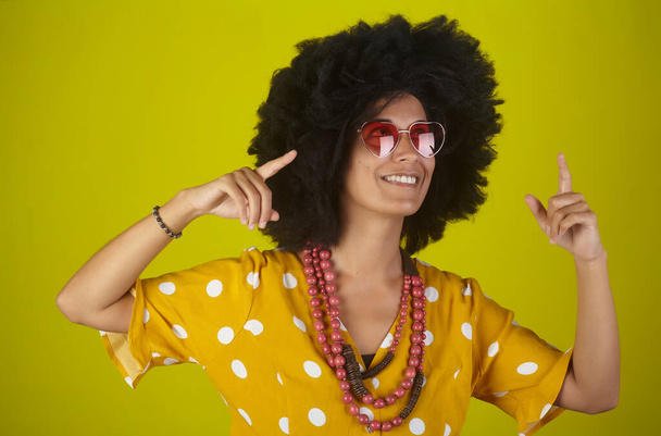 Young beautiful and smiling woman with curly afro hairstyle and heart shaped glasses over yellow background pointing and looking up, indicating direction with fingers. - Fotografie, Obrázek