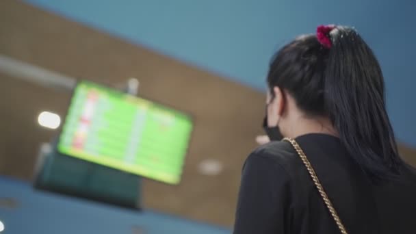 Pohled zezadu mladá žena nosí masky při pohledu na letištní obrazovku, terminálovou odletovou plošinu, veřejnou dopravu, čas nástupu, pandemii covid-19, novou normální společenskou vzdálenost, poslední hovor  - Záběry, video