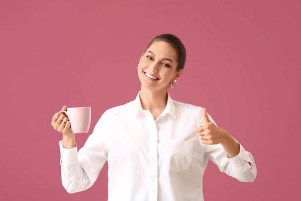 Beautiful young woman with cup of tea showing thumb-up on color background - 写真・画像