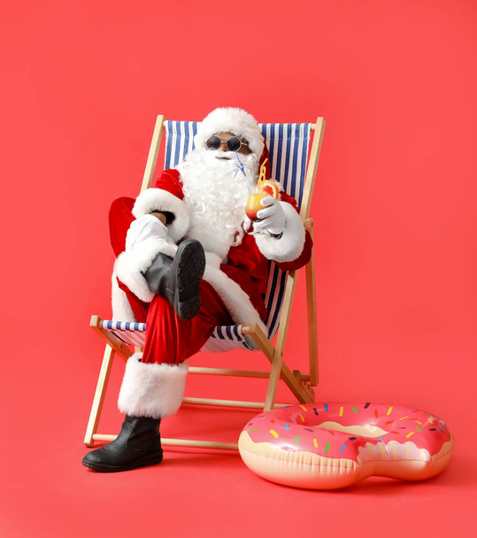 African-American Santa Claus with cocktail sitting on sun lounger against color background - Foto, Bild