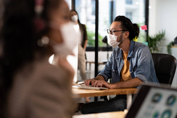 Asian Office employee with protective face mask working in new normal office with social distance practice to african colleague in foreground prevent coronavirus COVID-19 spreading. - Foto, imagen