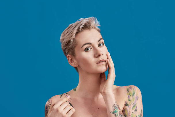 Portrait d'une jeune femme tatouée à moitié nue avec une peau parfaite regardant la caméra, posant avec la main sur son visage isolé sur fond bleu - Photo, image