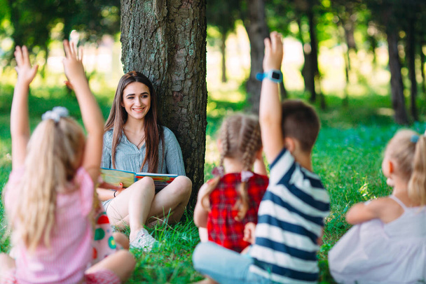 Children and education, young woman at work as educator reading book to boys and girls in park. - Photo, Image