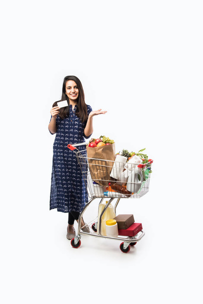 Indiai fiatal nő bevásárlókocsival vagy trolly tele élelmiszerekkel, zöldségekkel és gyümölcsökkel. Elszigetelt Teljes hosszúságú fotó fehér háttér - Fotó, kép