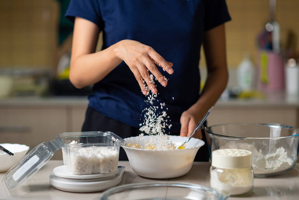 Frau fügt Kokosraspeln in Plätzchenteig hinzu, während sie zu Hause in der Küche hausgemachte Plätzchen zubereitet - Foto, Bild