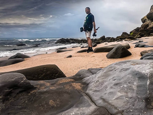 Мужчина фотографирует на скалистом пляже в черной футболке и белых шортах - Фото, изображение