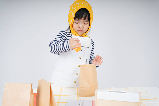 Nätti aasialainen thai lapsi pelata rooli tarjoilija yllään keltainen side pakkaus ruokaa paperipussi. - Valokuva, kuva