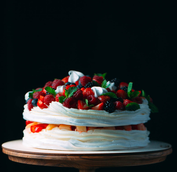Fruit cake. Cake decorated with berries on a wooden stand on a black background. - 写真・画像
