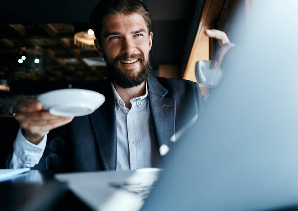 Επιχειρηματίας κάθεται σε ένα καφέ με ένα φλιτζάνι καφέ στα χέρια του μπροστά από ένα φορητό υπολογιστή επικοινωνίας τεχνολογίας - Φωτογραφία, εικόνα