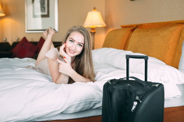 Smiling young businesswoman in bed staring at camera. Hotel room. the girl licks on the bed near the suitcase. - Photo, image