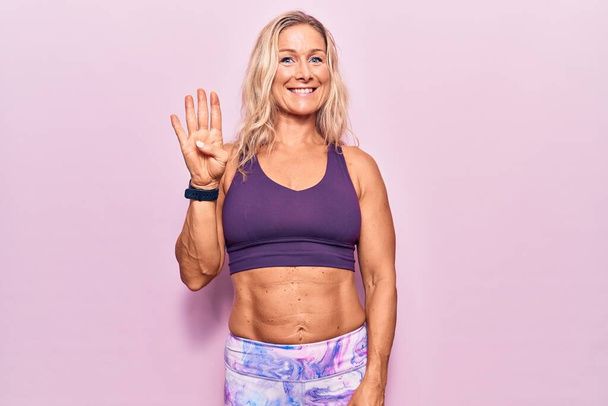 Kaukasische Blondine mittleren Alters trägt Sportbekleidung über rosa Hintergrund und zeigt mit Finger Nummer vier nach oben, während sie selbstbewusst und glücklich lächelt.  - Foto, Bild
