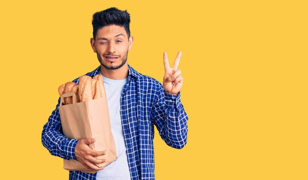 Beau jeune homme latino-américain tenant un sac en papier avec du pain souriant avec un clin d'œil heureux à la caméra faisant signe de victoire avec les doigts. numéro deux.  - Photo, image