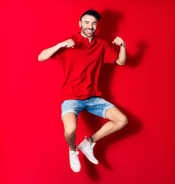 Young handsome deliveryman wearing cap smiling happy. Jumping with smile on face doing winner gesture with fists up over isolated red background - Foto, Imagem