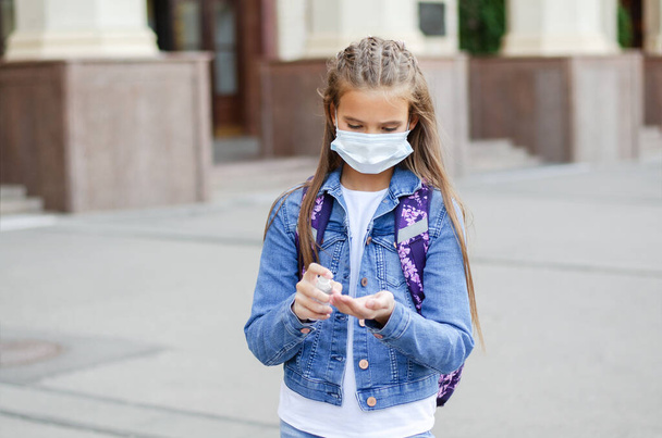 Little girl child with face mask and sanitizer. Education concept. Back to school after covid-19.  Preteen student disinfecting hands before school outdoors. - Фото, изображение