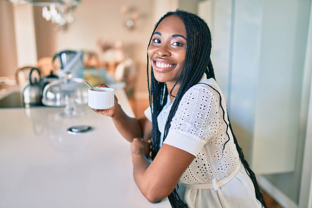 Joven afroamericana americana sonriendo feliz bebiendo taza de café en casa - Foto, Imagen