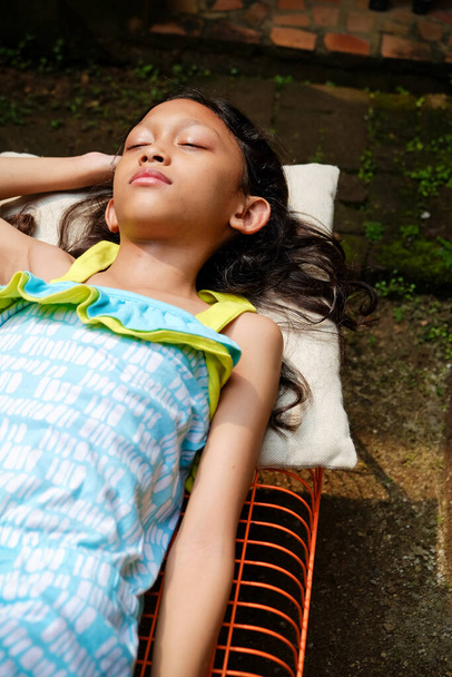 Χαριτωμένο κορίτσι ασιατικής εθνικότητας ξαπλωμένο σε ένα παγκάκι, κάνοντας ηλιοθεραπεία το πρωί στο σπίτι. το χέρι σηκώνεται για να καλύψει το πρόσωπό της από το σκληρό φως του ήλιου - Φωτογραφία, εικόνα