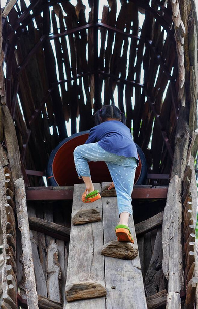 De achteraanzicht van een jong meisje klimmen op een houten steile helling in een park, grijpen de trappen met haar handen, zijn moedig maar voorzichtig. - Foto, afbeelding