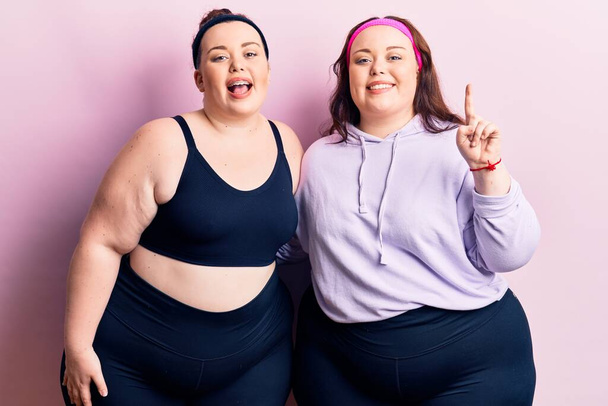 Young plus size twins wearing sportswear showing and pointing up with finger number one while smiling confident and happy.  - Photo, Image