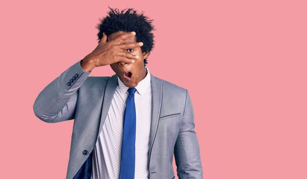 Knappe Afro-Amerikaanse man met afro haar draagt een zakelijke jas gluren in shock bedekken gezicht en ogen met de hand, kijkend door vingers met beschaamde uitdrukking.  - Foto, afbeelding