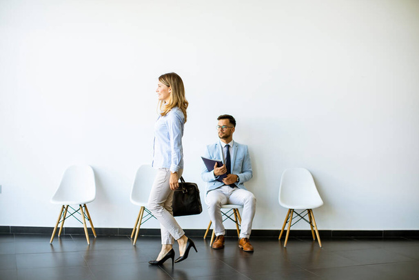 Femme d'affaires avec valise passant par un jeune homme assis à la chaise dans la salle d'attente avec un dossier en main avant une interview - Photo, image