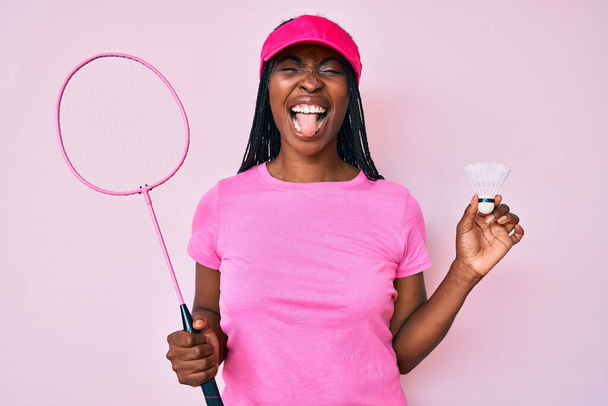 Afrykańska amerykanka z warkoczami trzymająca rakietę badmintona wystającą z języka szczęśliwa ze śmiesznym wyrazem twarzy.  - Zdjęcie, obraz